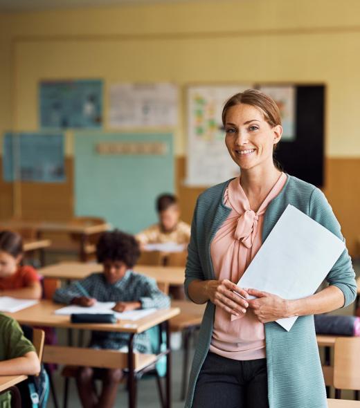 Happy teacher during a class at elementary school looking at camera. Her student are learning in the background.